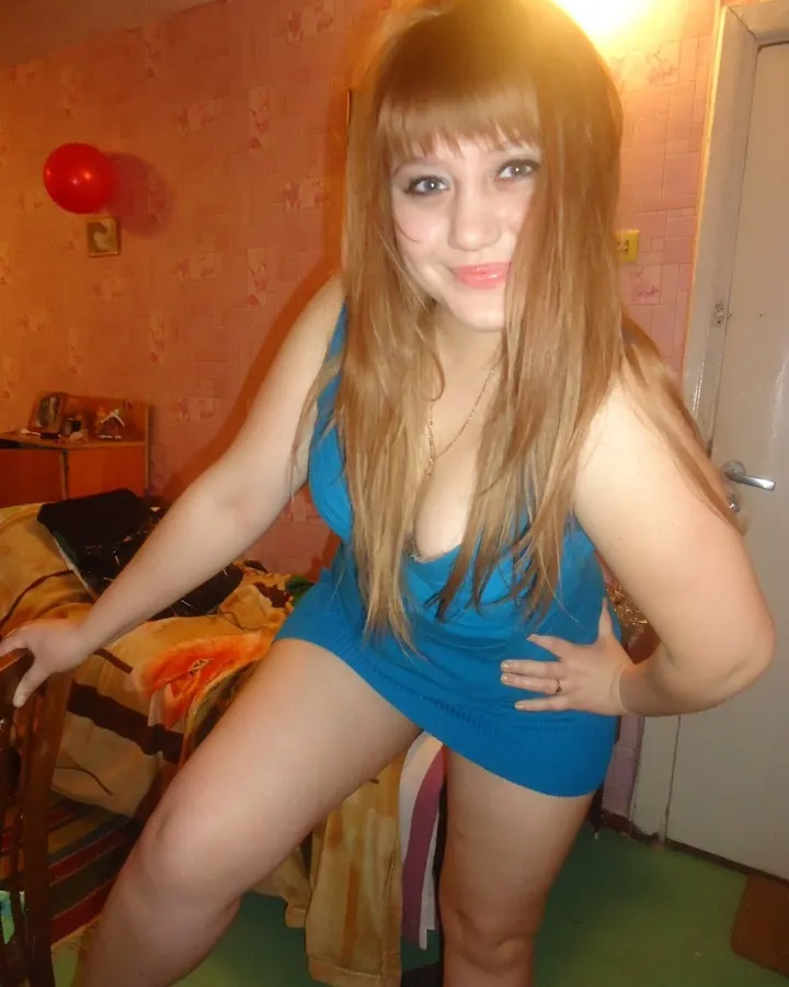 Jeune femme aux cheveux blonds portant une robe bleue courte, souriante, dans une chambre avec un ballon rouge en arrière-plan.