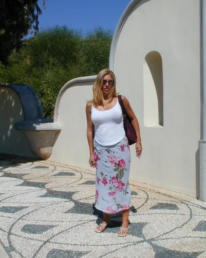Femme blonde aux cheveux courts, portant un haut blanc et une jupe fleurie, posant sur un trottoir décoré de mosaïques, avec un sac à dos violet.