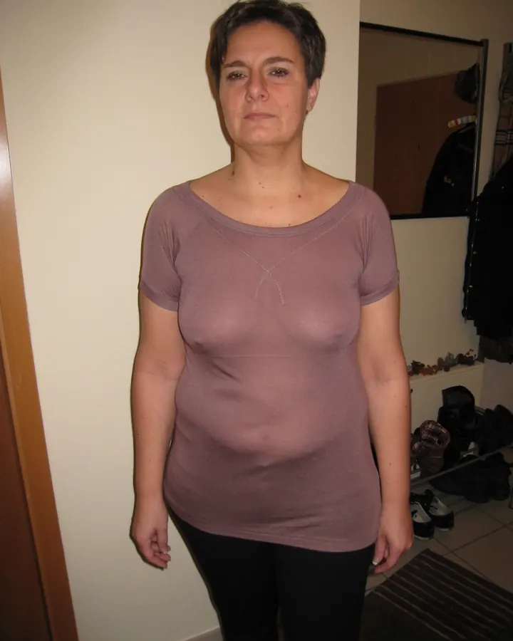 Femme aux cheveux bruns, portant un haut marron, posant debout dans une pièce, avec une expression neutre.
