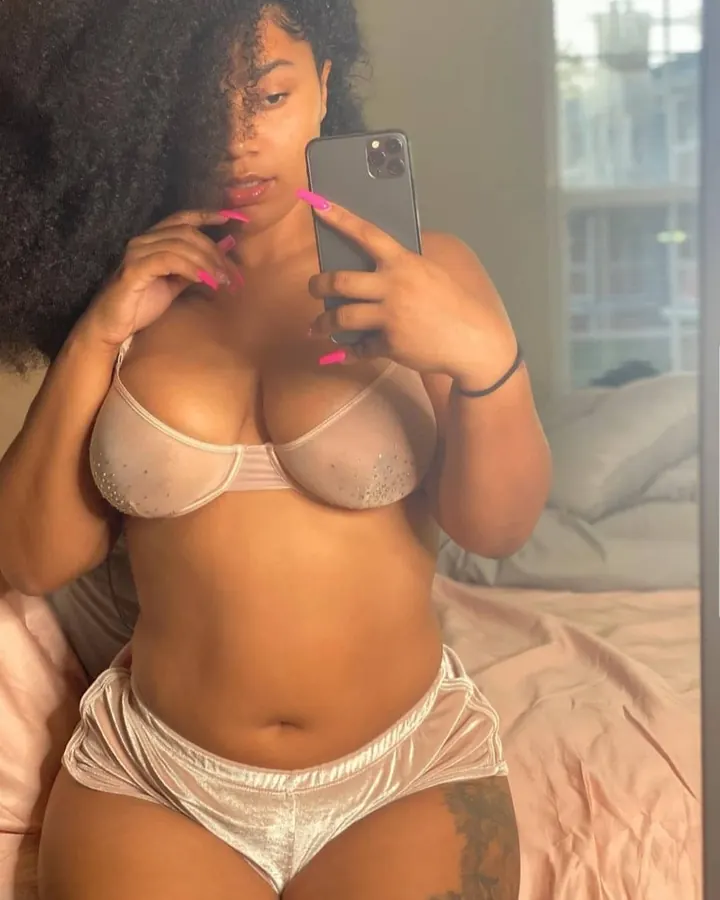 Jeune femme africaine aux cheveux noirs bouclés, portant un soutien-gorge beige et un short assorti, prenant un selfie dans un miroir, avec un tatouage sur la cuisse.