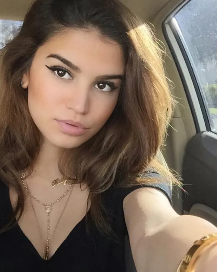 Jeune femme latino aux cheveux châtains, maquillée avec des yeux soulignés, prenant un selfie dans une voiture, portant un haut noir et des bijoux dorés.
