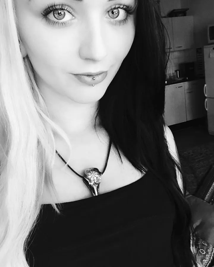 Photo en noir et blanc d'une femme aux cheveux foncés et blonds, portant un collier avec un pendentif, regardant l'appareil.