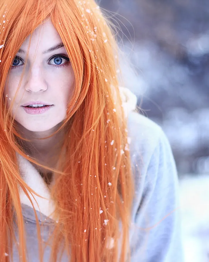 Femme aux cheveux rouges, en pull gris, dans un paysage enneigé, avec des flocons sur les cheveux.