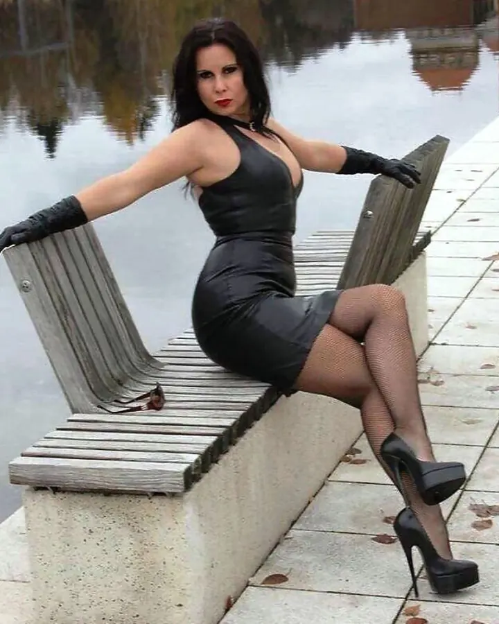 Femme aux cheveux bruns-noirs, portant une robe noire en cuir, des gants noirs et des chaussures à talons hauts, assise sur un banc en bois près d’un plan d’eau.
