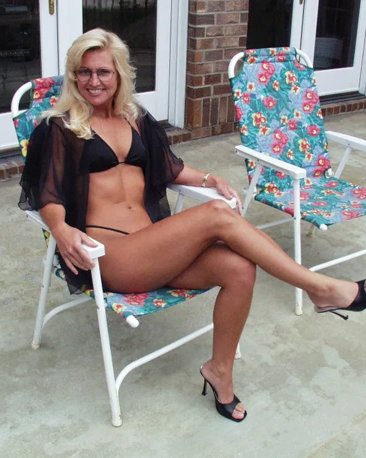 Femme aux cheveux blancs, portant un haut noir et un short en jean, assise sur une chaise de jardin, les jambes croisées, les mains sur les genoux.